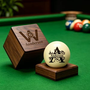 May include: A dark brown wooden box with a lid featuring a laser-engraved letter W with an 8-ball graphic and the text Annderson. Beside it, a white billiard ball sits on a wooden base, engraved with a large letter A and the name Anderson.