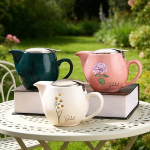 May include: Three ceramic teapots in teal, pink, and cream, each with a stainless steel lid. The teapots feature floral designs and the name "Rachel" in script. They are displayed on a white table with a green background.