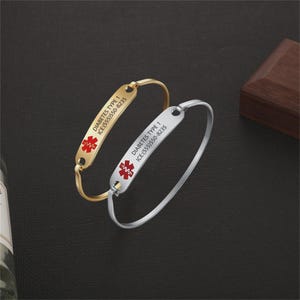 Puede incluir: Dos pulseras de alerta médica, una dorada y otra plateada, con el texto "DIABETES TYPE 1" y un número de contacto de emergencia. Cada pulsera tiene un símbolo médico rojo.