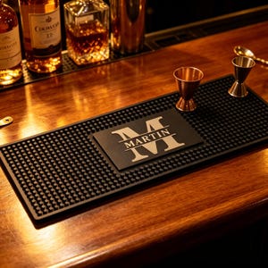 Puede incluir: Una alfombrilla de bar rectangular de goma negra con una superficie texturizada de puntos. El centro presenta un panel rectangular elevado que muestra una letra M grande con el nombre MARTIN impreso horizontalmente en beige claro.