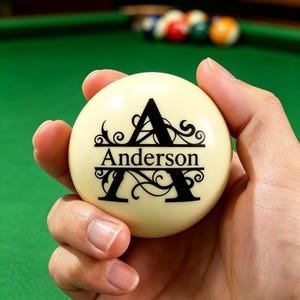 May include: A cream-coloured billiard ball featuring a black decorative letter A with the name Anderson printed in the centre. The design includes intricate scrollwork accents surrounding the letter and the name on the smooth, round surface.