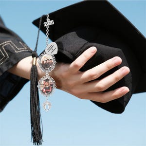 May include: A graduation tassel charm with a silver chain and photo frames. The charm features a '2026' pendant, a heart-shaped charm with text, and two oval frames holding photos. A black graduation cap is held in the background.