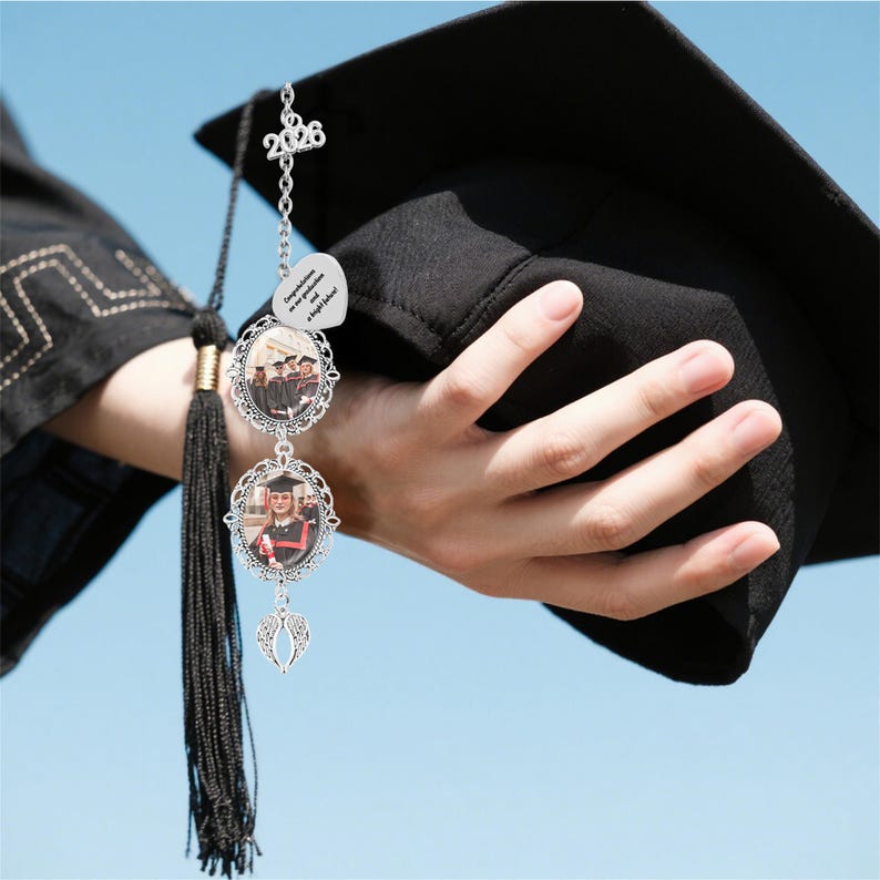 May include: A graduation tassel charm with a silver chain and photo frames. The charm features a '2026' pendant, a heart-shaped charm with text, and two oval frames holding photos. A black graduation cap is held in the background.