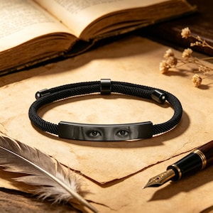 May include: A black cord bracelet featuring a central metal bar engraved with a detailed grayscale image of a pair of human eyes. The bracelet includes a sliding adjustment bead and two cylindrical end caps, all finished in a matte black tone.