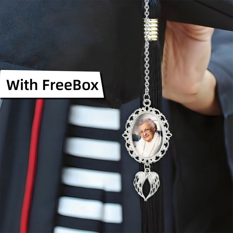 May include: A graduation tassel charm with a silver frame holding a photo of an elderly woman. Below the photo is a silver heart with angel wings. The charm hangs from a black tassel and is attached to a graduation cap.