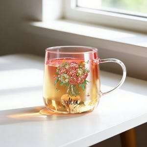 Könnte beinhalten: Eine transparente Glas-Tasse, gefüllt mit einer hellen Flüssigkeit, mit einem Blumendesign und dem Namen "Alex" auf einem herzförmigen Anhänger. Die Tasse hat einen gebogenen Griff und steht auf einer weißen Oberfläche in der Nähe eines Fensters.