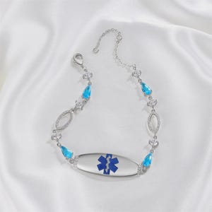 May include: A silver-toned medical alert bracelet with a blue Star of Life symbol. The bracelet features teardrop-shaped blue gemstones and oval-shaped clear stones. The bracelet is displayed on a white, silky surface.