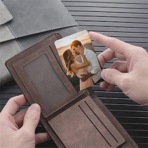 May include: Brown bi-fold wallet with a clear ID window and card slots. A small photo card featuring a couple kissing is held above the wallet. A gray leather-like accessory is in the background.