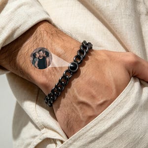 Può includere: Un bracciale a catena gourmette in metallo nero con un ciondolo circolare centrale. Il ciondolo proietta una fotografia circolare in bianco e nero che mostra una persona baciata su entrambe le guance da due bambini, creando un effetto visivo.
