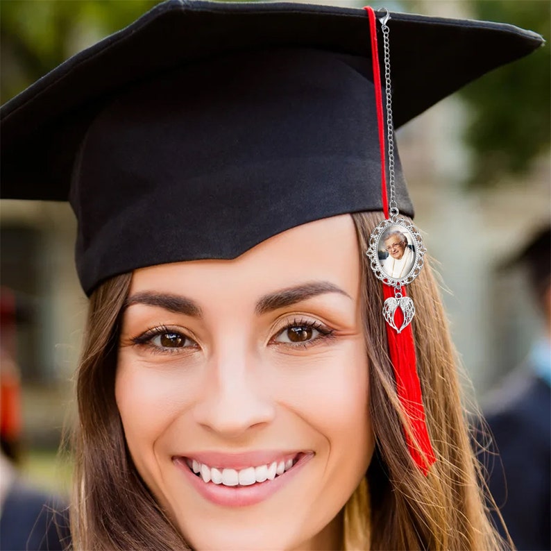 May include: Black graduation cap with a red tassel and a decorative charm. The charm features a small framed photo and a silver-colored heart with wings. The woman in the photo is smiling.