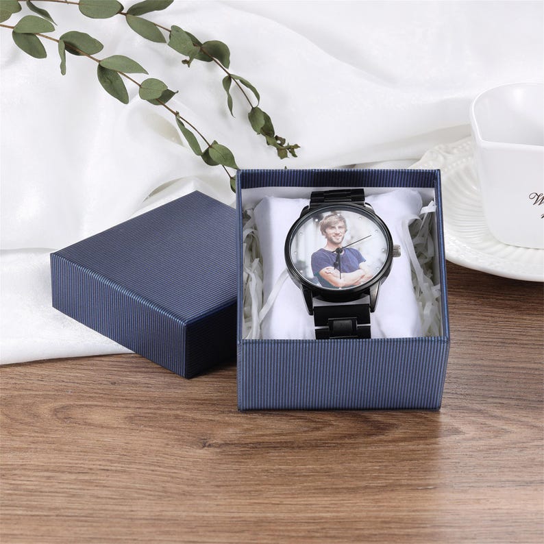May include: A black wristwatch with a custom photo dial featuring a person wearing a blue shirt, presented in a dark blue textured gift box with a white cushion. The watch has a black metal link band and a round face with simple black hands.