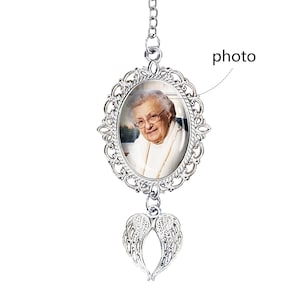 May include: A silver-colored memorial photo pendant with an oval frame and a picture of a smiling elderly woman. The pendant has a chain and a pair of angel wings at the bottom. The word "photo" is written on the image.