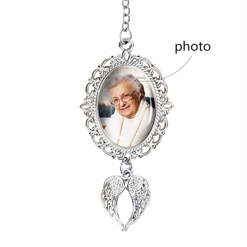May include: A silver-colored memorial photo pendant with an oval frame and a picture of a smiling elderly woman. The pendant has a chain and a pair of angel wings at the bottom. The word "photo" is written on the image.