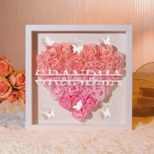 May include: A framed artwork featuring a heart-shaped arrangement of pink roses. The word "GRANDMA" is displayed in white, along with the phrase "we love you." White butterflies and names are also included.