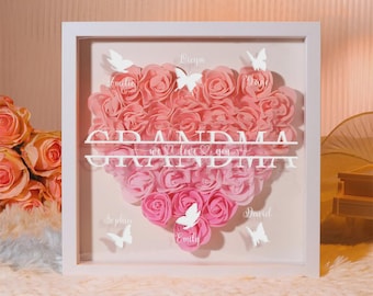 Può includere: Un'opera d'arte incorniciata con un cuore di rose rosa. Il testo bianco recita "NONNA" e "ti amiamo". Sono incluse anche farfalle bianche e nomi. L'opera d'arte è in una cornice bianca.