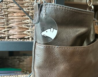 May include: A brown leather shoulder bag with a silver chain and a clear charm featuring a black and white rat illustration.