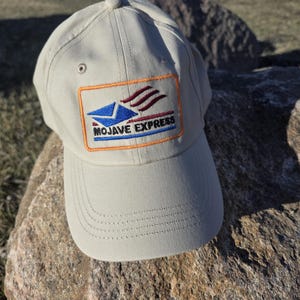 May include: A beige baseball cap featuring an embroidered rectangular patch on the front. The patch displays a blue envelope icon with red motion lines and the text MOJAVE EXPRESS in black, all outlined in an orange border with stitching.