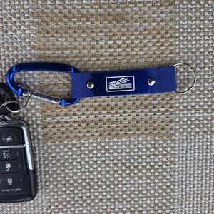 May include: A blue keychain with a carabiner clip and a key ring. The keychain has a blue fabric strap with the "Mojave Express" logo printed on it. A car key fob is visible on the left.
