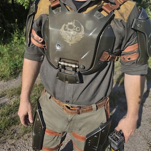 Puede incluir: Una armadura de pecho de color verde oliva oscuro con un emblema central de un engranaje y una espada, sobre una camisa gris oscuro. La armadura incluye hombreras y protectores de muslo a juego sujetos con correas de cuero marrón y hebillas.