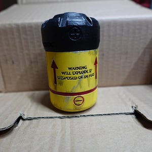 May include: A cylindrical object with a black top and yellow body. The yellow section has red arrows and text that reads "WARNING WILL EXPLODE IF DISPOSED OF IN FIRE". The object is inside a cardboard box.