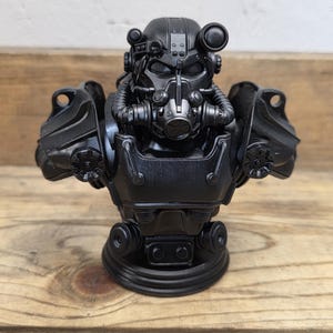May include: A black bust of a power armor helmet, featuring intricate details like a respirator, multiple lenses, and shoulder armor. The helmet is set on a wooden surface.