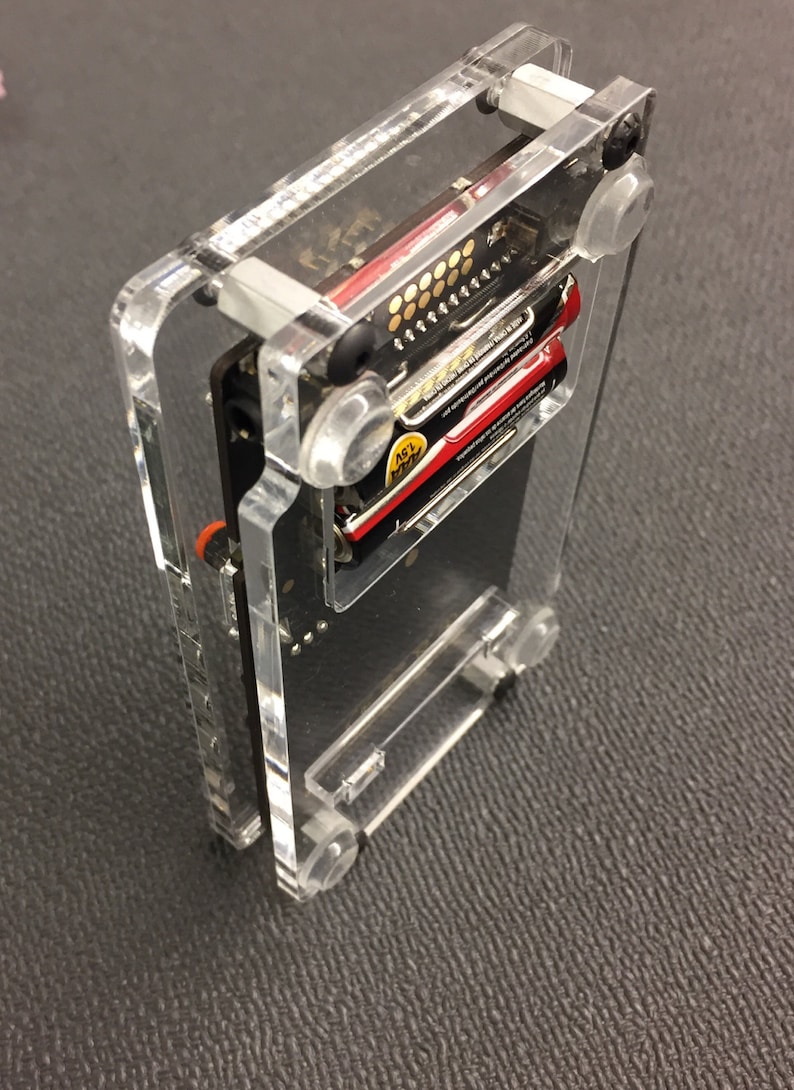 May include: A clear acrylic case with a black circuit board inside. The case has two AA batteries inside and clear plastic standoffs.