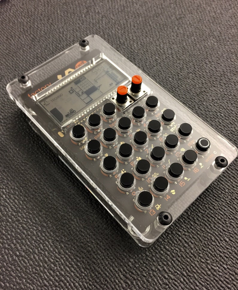 May include: A clear plastic case with a black electronic device inside. The device has a small LCD screen, several black buttons, and two orange knobs. The text "factory" is visible on the device.