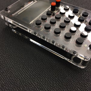May include: A clear plastic case with a black and white grid of buttons. The buttons have red and black knobs. There is a small screen in the top left corner of the case.