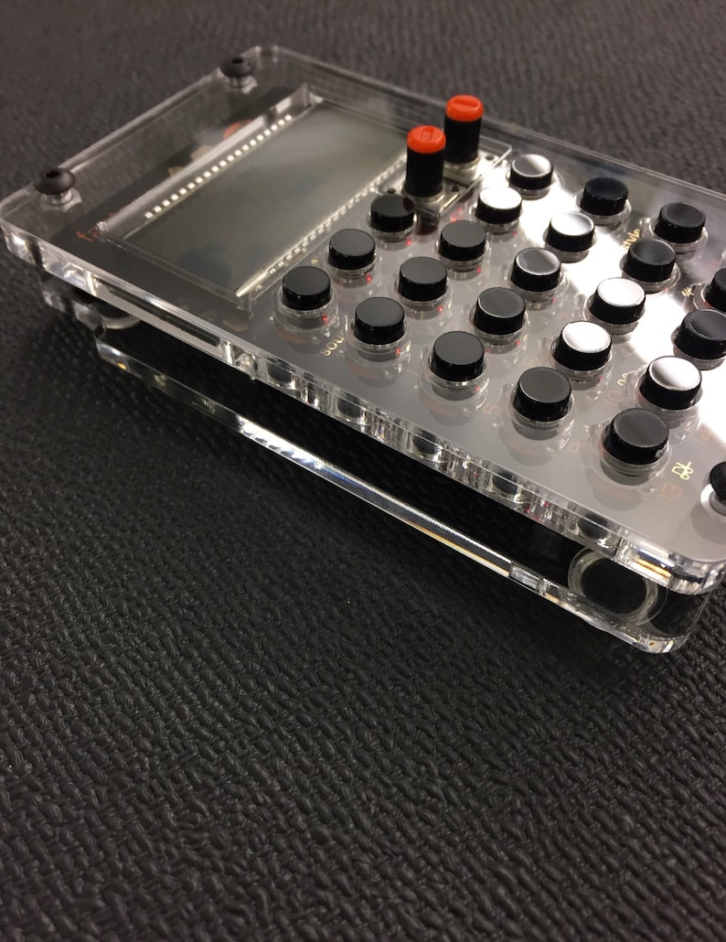 May include: A clear plastic case with a black and white grid of buttons. The buttons have red and black knobs. There is a small screen in the top left corner of the case.