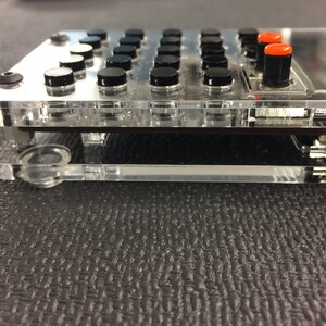 May include: A clear plastic case with a black and red electronic device inside. The device has a grid of black buttons and a small screen.