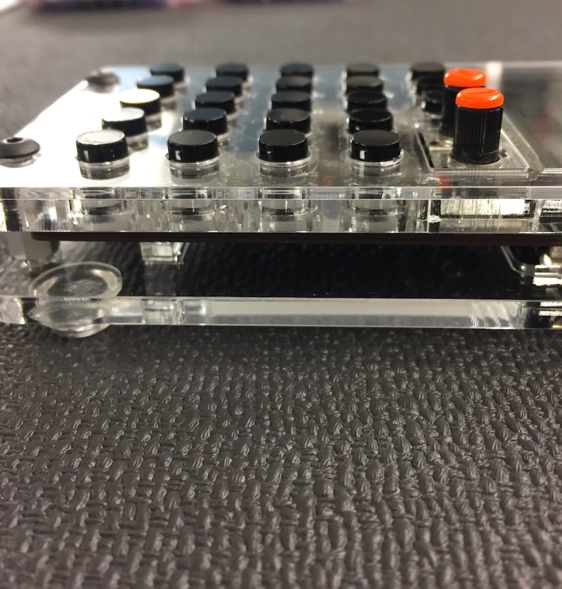 May include: A clear plastic case with a black and red electronic device inside. The device has a grid of black buttons and a small screen.