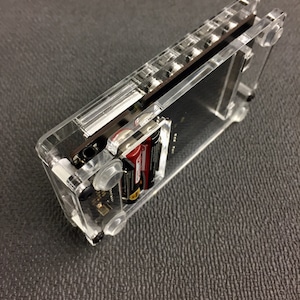 May include: A clear plastic case with a black circuit board inside. The case has a slot for batteries and a clear window on the front. The case is sitting on a gray surface.