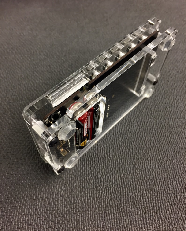 May include: A clear plastic case with a black circuit board inside. The case has a slot for batteries and a clear window on the front. The case is sitting on a gray surface.
