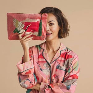 Puede incluir: Un clutch de terciopelo rosa con un borla verde y un diseño de grúa bordado. El clutch se sostiene frente a una persona que lleva una camisa estampada en rosa y blanco.