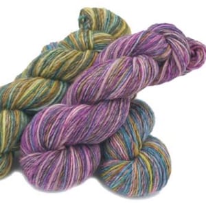 May include: Two skeins of colorful yarn. One skein is a mix of greens, yellows, and browns. The other is a blend of purples, pinks, and blues. The yarn is thick and appears to be hand-dyed, perfect for knitting or crocheting projects.