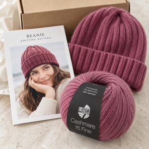 May include: A dark pink ribbed beanie, a ball of matching yarn with a label reading 10% Polyamide, 10% Merino Wool, 80% Cashmere, Lana Grossa Cashmere 16 Fine, and a printed knitting pattern booklet titled Beanie Knitting Pattern.