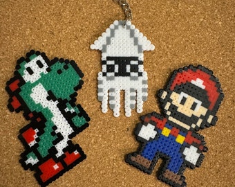 May include: Three pixel art keychains featuring characters from popular video games. The first keychain is a green Yoshi, the second is a white squid, and the third is a red and blue Mario.