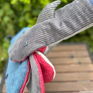 Pode incluir: Um conjunto de três luvas de forno em forma de tubarão. As luvas são feitas de tecido de veludo cotelê cinza, azul e vermelho. A luva cinza tem uma barbatana e olhos de botão. O interior das luvas é forrado com um padrão floral vermelho e branco.