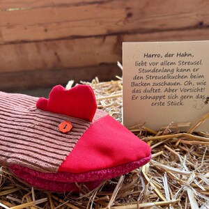 Könnte beinhalten: Ein kissenförmiges Huhn mit rotem Kamm und Schnabel sowie einem braun-rosa gestreiften Körper. Eine kleine Karte mit deutschem Text ist auf dem Stroh aufgestellt. Das Kissen liegt auf einem Strohbett.