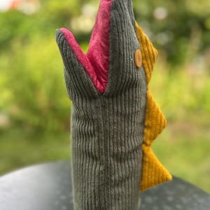 May include: A novelty bottle sleeve in the shape of a sea monster. The olive green corduroy sleeve has a red patterned interior, yellow fin, and a wooden button. The base is tan. Designed to fit over a standard bottle, it is a fun accessory.