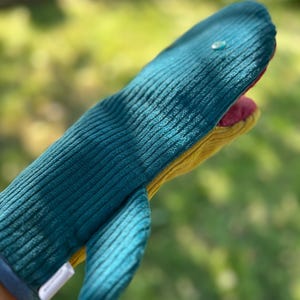 Puede incluir: Un guante de cocina con forma de ballena hecho de tela de pana color verde azulado con textura acanalada. El guante presenta un pequeño ojo circular azul claro, una parte inferior color amarillo mostaza y un forro interior rosa brillante.