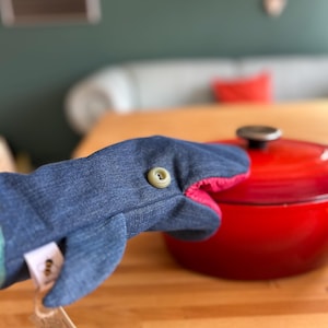 Puede incluir: Un guante de cocina con forma de tiburón de mezclilla con un botón verde y un interior rosa. El guante está junto a una olla roja con un asa negra. Una etiqueta está adherida al guante.