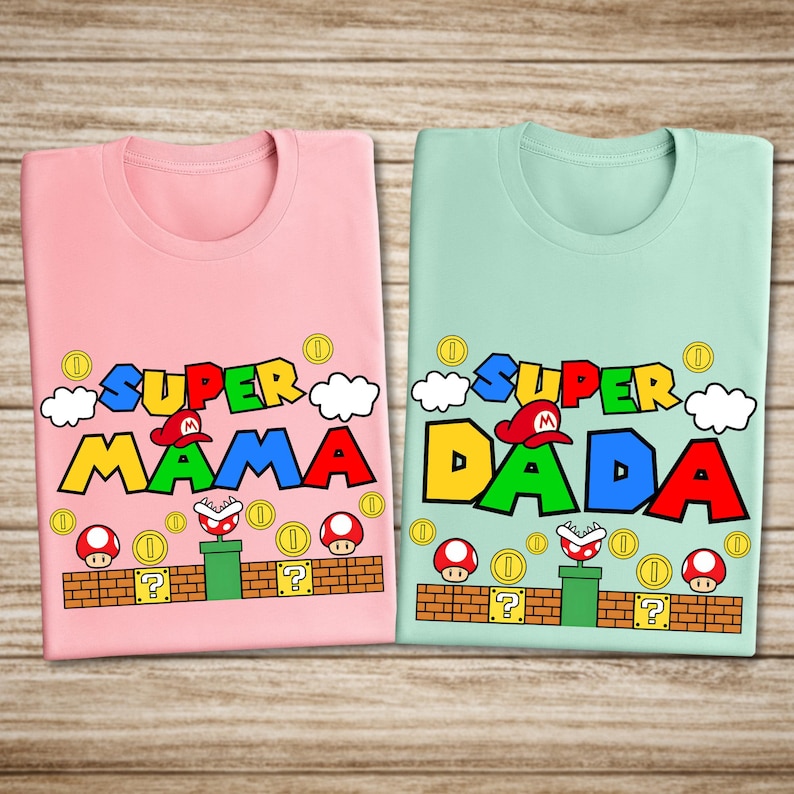 two t-shirts with a Mario-themed design, specifically the Super Mario Bros. video game character. The t-shirts are pink and green, and they display the text "SUPER MAMA" and "SUPER DADA" along with Mario mushroom graphics and other Mario-related elements.