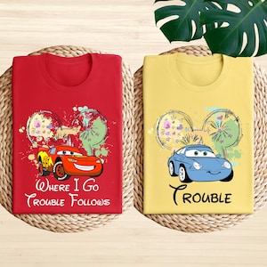 two t-shirts with cartoon car designs and text, laid out on a woven mat.
