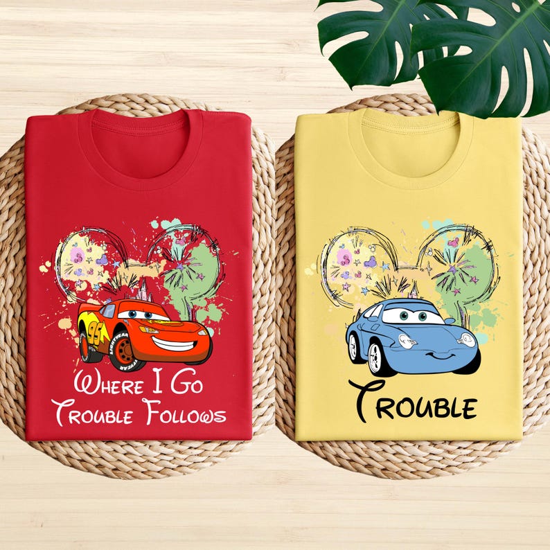 two t-shirts with cartoon car designs and text, laid out on a woven mat.