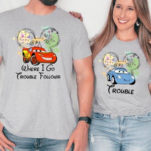 A man and a woman are standing next to each other, both wearing gray t-shirts with a cartoon car and the text "Where I go, trouble follows" printed on them.