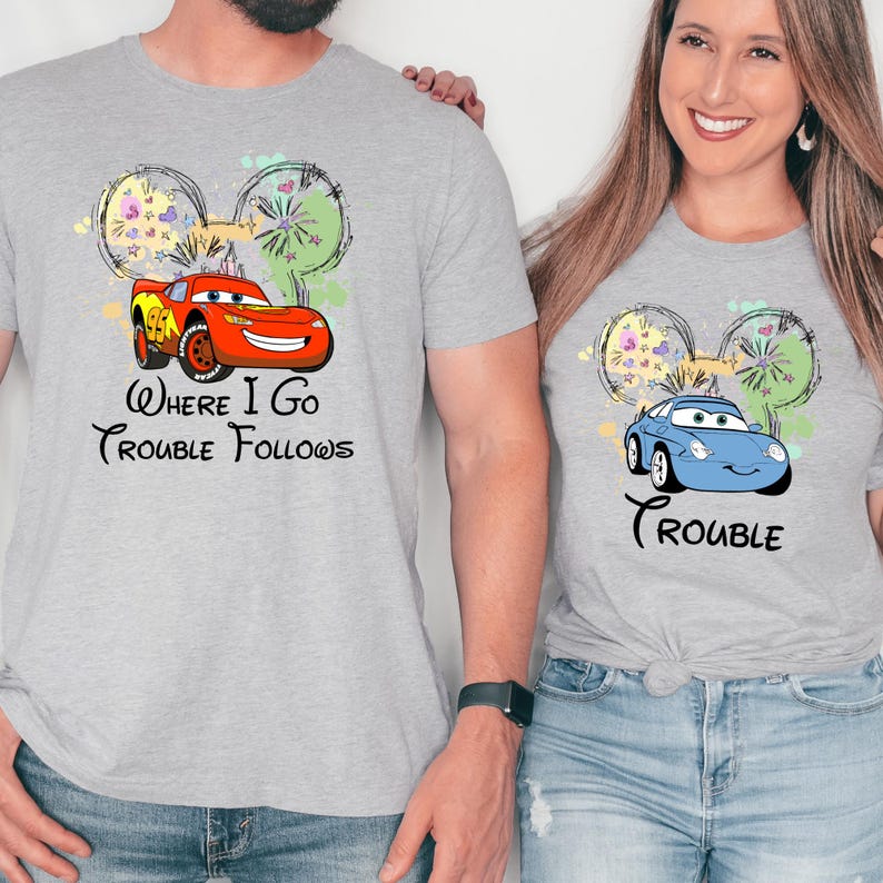 A man and a woman are standing next to each other, both wearing gray t-shirts with a cartoon car and the text "Where I go, trouble follows" printed on them.