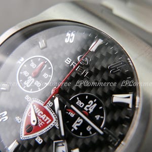 Op de afbeelding: Close-up van een zilveren polshorloge met een zwarte carbon fiber wijzerplaat. Het horloge heeft meerdere sub-wijzerplaten, waaronder een chronograaf en een 24-uurs indicator. De merknaam "BREIL" is zichtbaar, samen met het "DUCATI" logo. De band is van roestvrij staal.