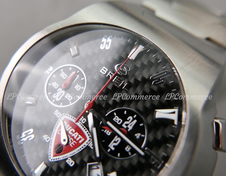 Op de afbeelding: Close-up van een zilveren polshorloge met een zwarte carbon fiber wijzerplaat. Het horloge heeft meerdere sub-wijzerplaten, waaronder een chronograaf en een 24-uurs indicator. De merknaam "BREIL" is zichtbaar, samen met het "DUCATI" logo. De band is van roestvrij staal.