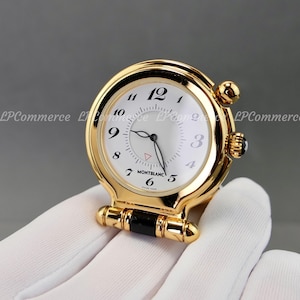 Könnte beinhalten: Eine goldfarbene Taschenuhr mit weißem Zifferblatt, schwarzen arabischen Ziffern, einem zentralen Hilfszifferblatt und dem Montblanc-Logo. Die Uhr hat ein poliertes rundes Gehäuse, eine seitliche Krone mit schwarzem Akzent und einen Klappfuß.